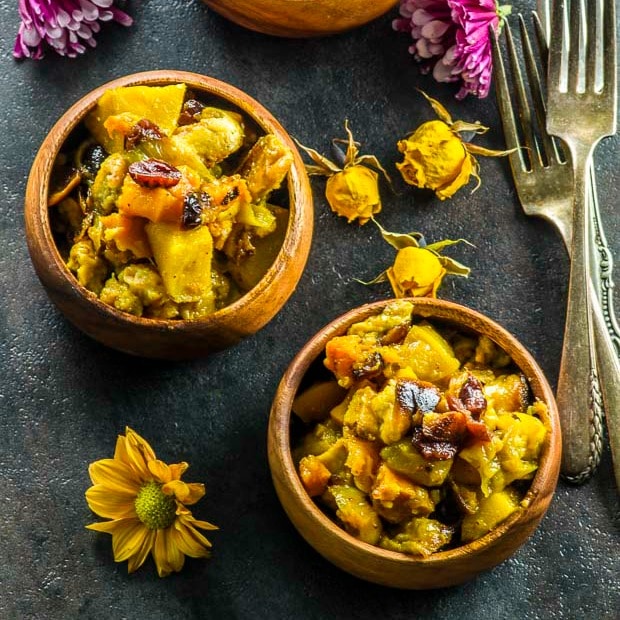 Birds eye view of the vegan butternut squash apple stuffing served in small wood bowls on a dark gray surface, with two forks placed on the right hand side and pink and yellow flowers scattered