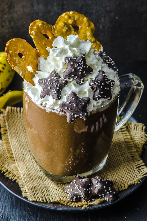 Close up of a glass mug of peanut butter and banana vegan hot chocolate topped with coconut whipped cream, three pretzel thins and 5 chocolate coversed star shaped cookies, on a small plate with two woven coasters