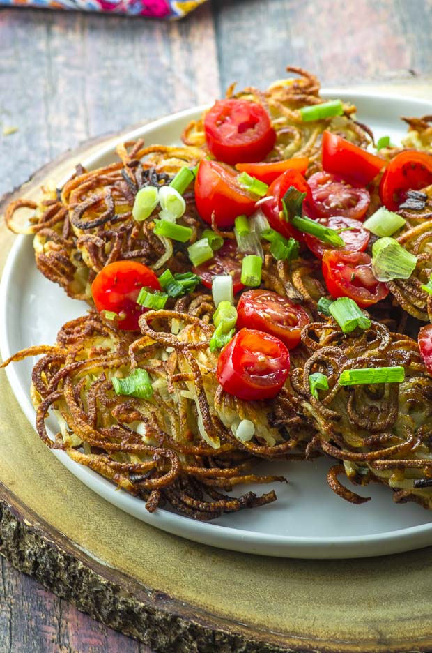 Potato latkes topped with sliced grape tomatoes and chopped scallions on a white plate, on top of a round wooden board
