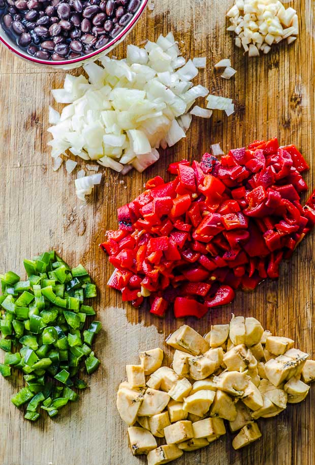 Diced roasted red peppers, jalapeno peppers, onions, plantains and garlic, and a bowl of black beans on a wooden cutting board