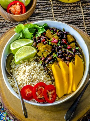 A large white bowl with black beans and rice with plantains, topped with mango slices, two lime slices, hot pepper slices and cilantro
