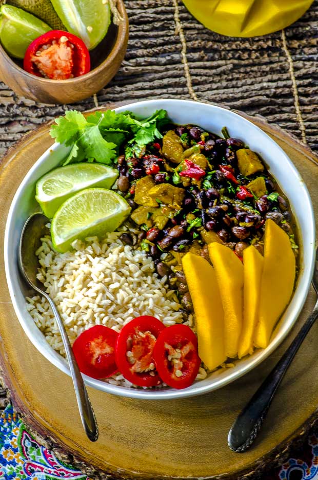 A large white bowl with black beans and rice with plantains, topped with mango slices, two lime slices, hot pepper slices and cilantro