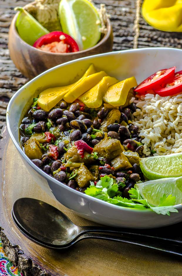 A large white bowl with black beans and rice with plantains, topped with mango slices, two lime slices, hot pepper slices and cilantro on a round wooden board, wit htwo silver spoons on the left side, and a small wooden board with lime wedges and a pepper slice in it