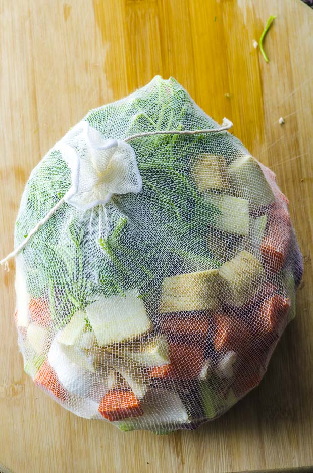 A tied up cheese cloth bag with cut carrots, celery, parsnips , red onions, yellow onion, shiitake mushroom stems, dill, parsley, and a whole head of garlic cut in half
