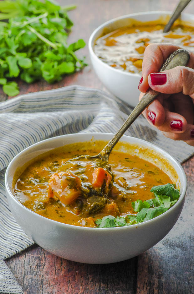 Side view of a bowl of peanut soup with spinach and sweet potatoes. A. hand is shown taking a spoonful of peanut soup from the bowl . in the background there is another white bowl of soup and a bunch of cilantro.