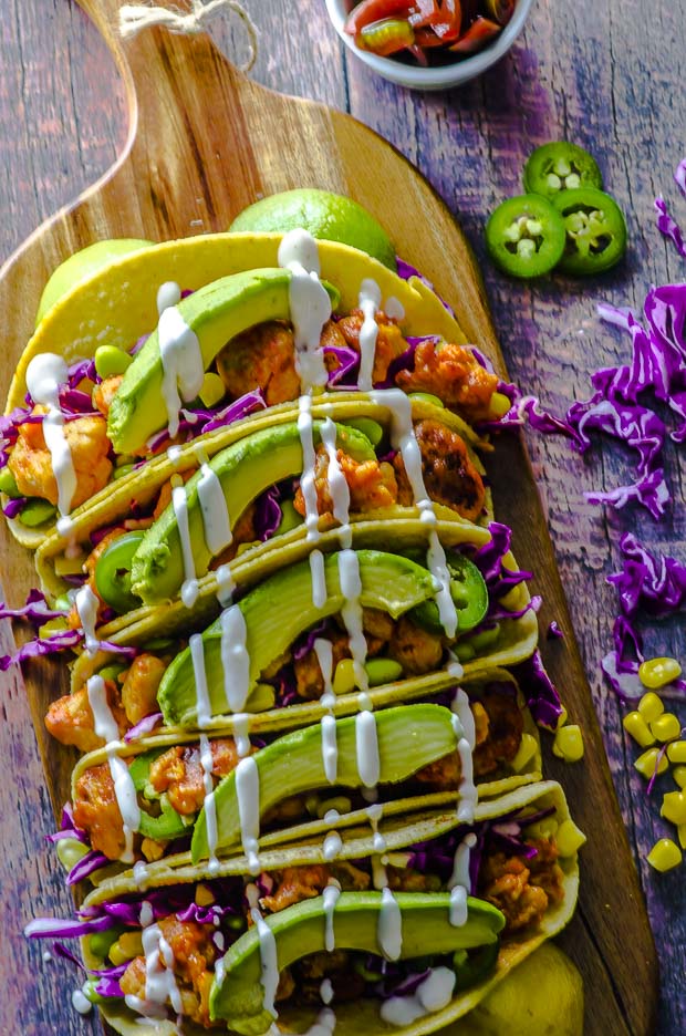 Birds eye view of five buffalo cauliflower vegan tacos with shredded purple cabbage, edamame and corn, topped with avocado slices and cashew cream, on a wooden board. On the right side ther are 3 jalapeno pepper slices, some shredded cabbage and a few corn kernels