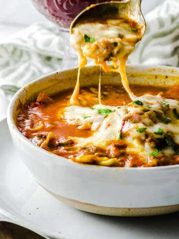 Close up of the lasagna soup with melted cheese on top and a silver spoon scooping it put, in a white bowl over  a white surface