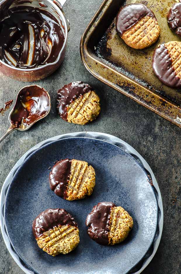 Recipes with dates:: Birds eye view of three almond butter cookies on a small blue plate, one off the plate with a silver spoon coated with melted chocolate and a small saucepan with melted chocolate on the left, and part of a cookie sheet with three cookies on it on the right