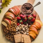 a round wood board with a bowl with chocolate hummus next to strawberries, apples, peaches, pistachios, pretzels and graham crackers.
