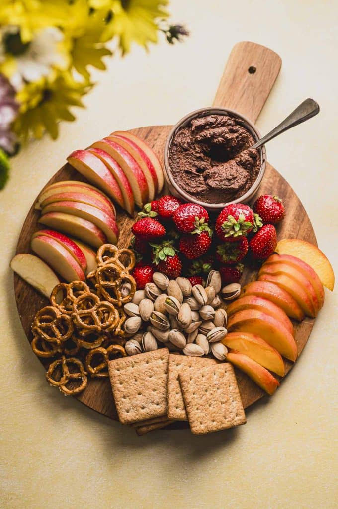 a round wood board with a bowl with chocolate hummus next to strawberries, apples, peaches, pistachios, pretzels and graham crackers.