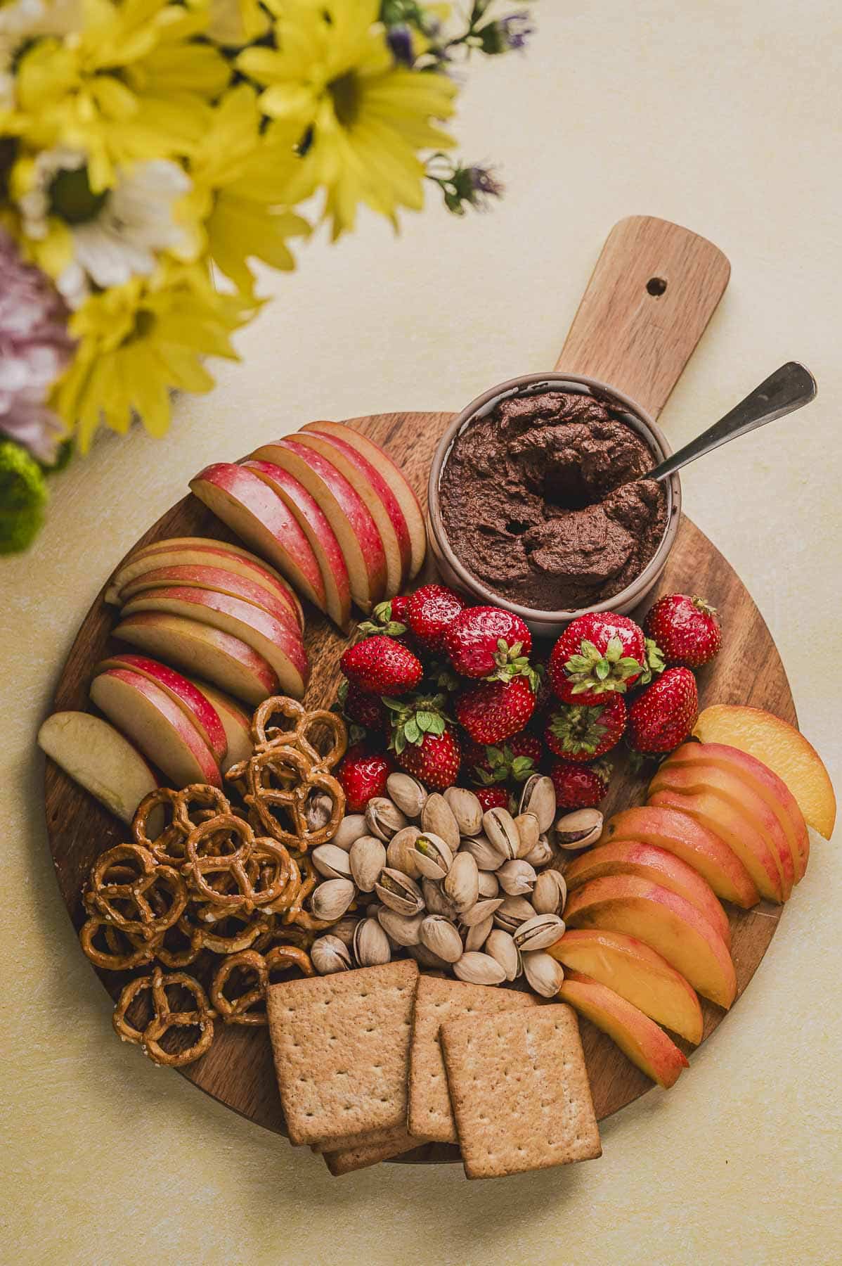 a round wood board with a bowl with chocolate hummus next to fruit, nusts, crakers and pretzels.