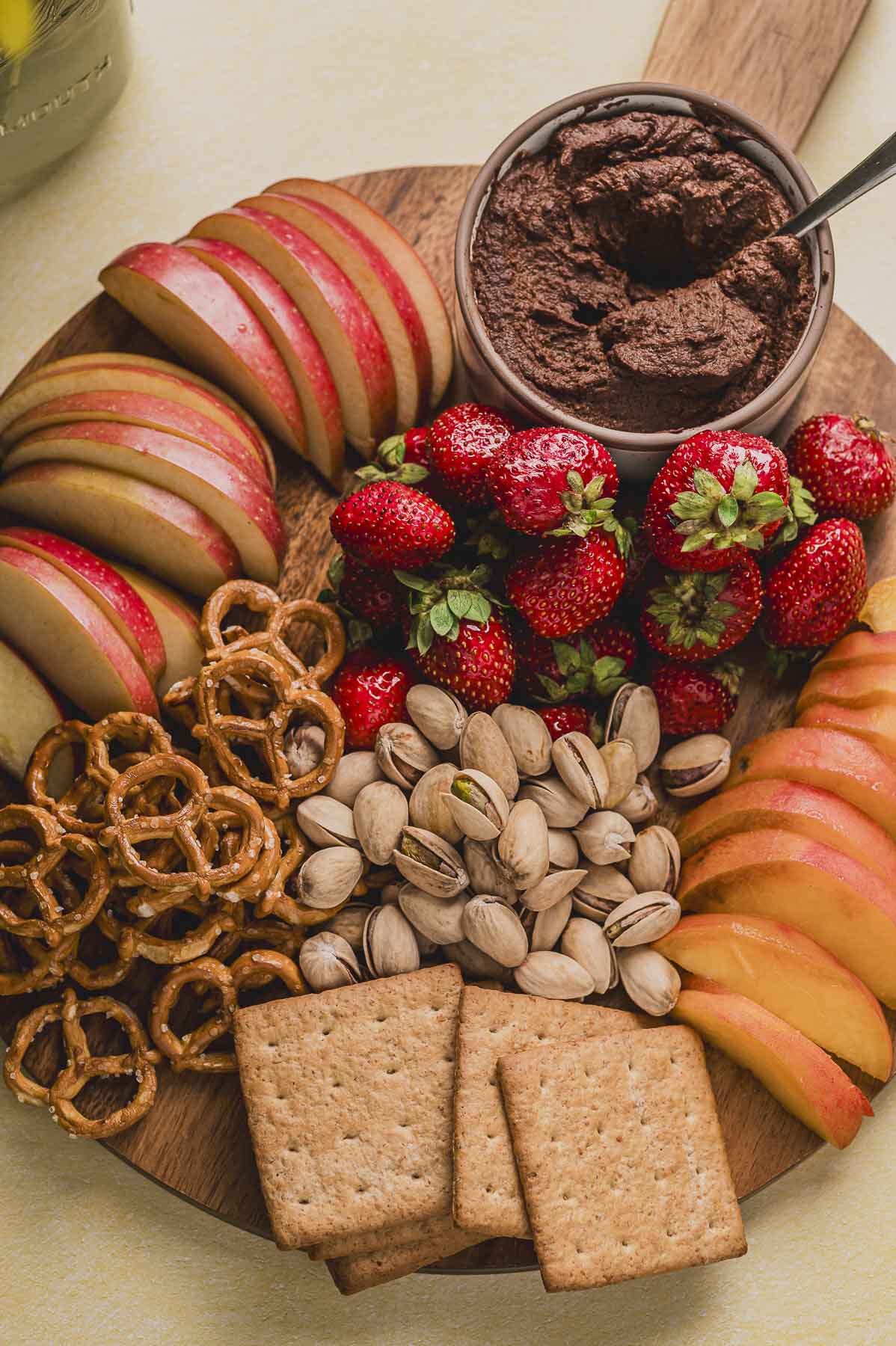 Close up view of a round wood board with a bowl with chocolate hummus next to strawberries, apples, peaches, pistachios, pretzels and graham crackers.