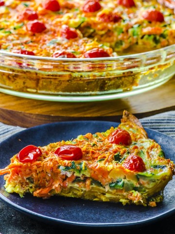 Close up of a slice of quiche with sweet potato crust on a small dark blue plate. In the background there a clear baking dish with the rest of the quiche