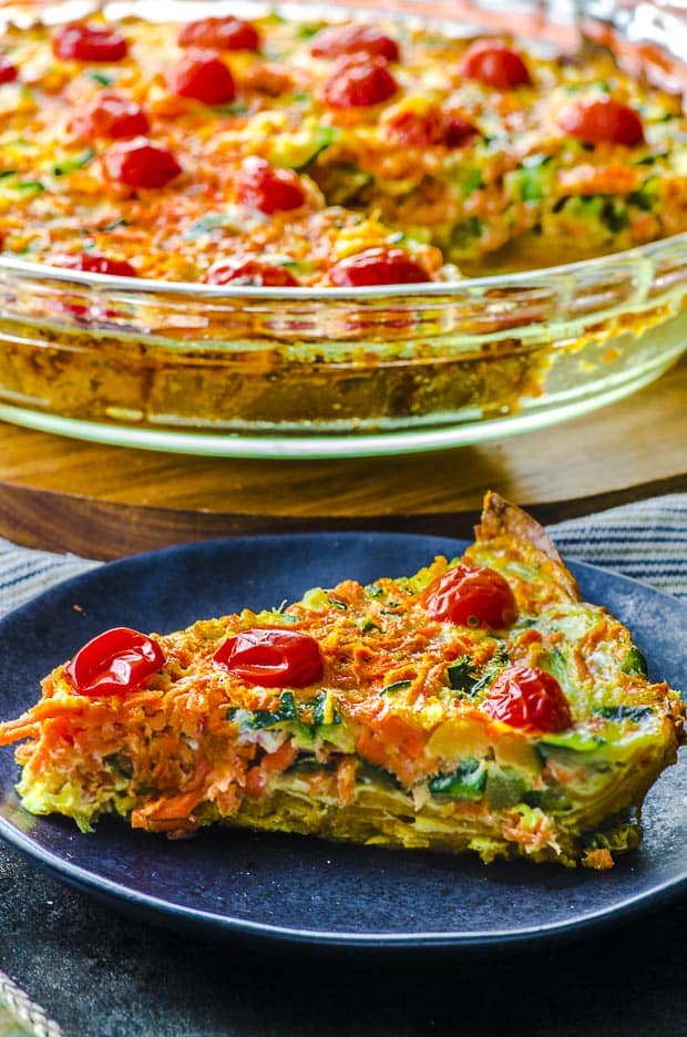 Nahaufnahme einer Scheibe Quiche mit Süßkartoffelkruste auf einem kleinen dunkelblauen Teller. Im Hintergrund eine klare Auflaufform mit dem Rest der Quiche. Ein vegetarisches Pessach-Rezept.