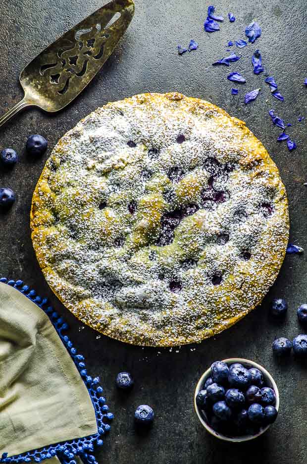 Birds eye view of the lemon blueberry cake, a silver cake cutter, a small bowl of bluerries, a cream color cloth napkin with blue fringes, and some scattered blueberries on a dark gray surface