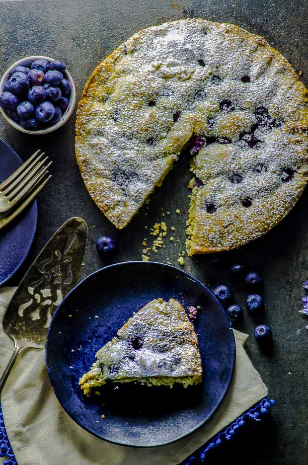 Birds eye view of a slice of the lemon blueberry cake, on a a cream color cloth napkin with blue fringes, a silver cake cutter, a small bowl of bluerries, and some scattered blueberries on a dark gray surface