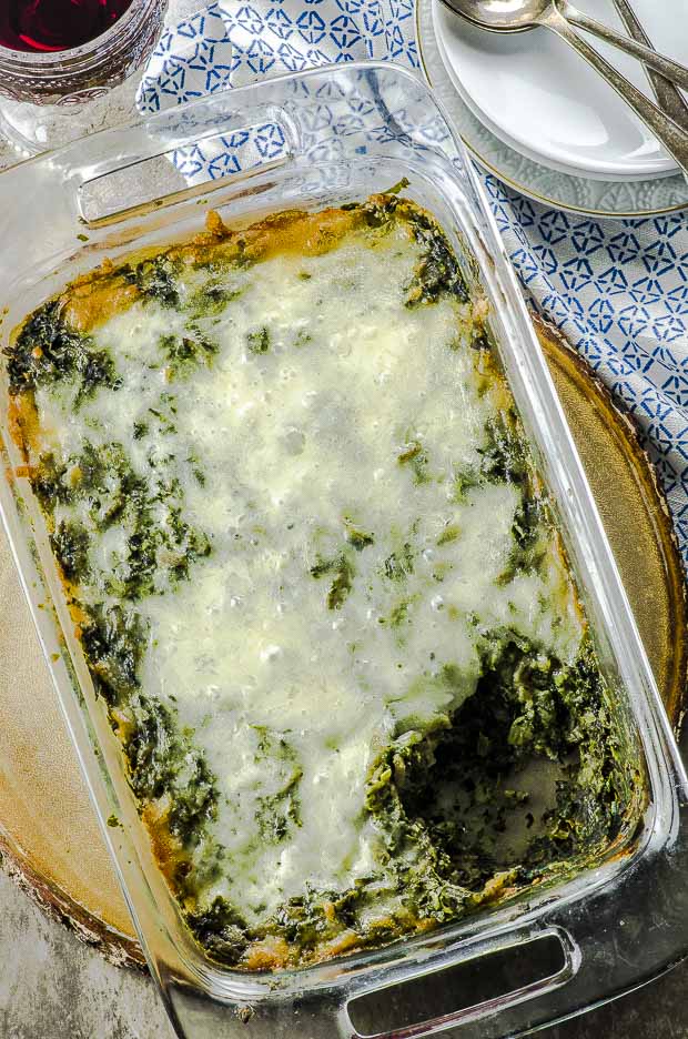 Birds eye view of high protein spinach pasta bake in a rectangular clear baking dish, on a round wooden board on a blue and white patterned cloth napkin
