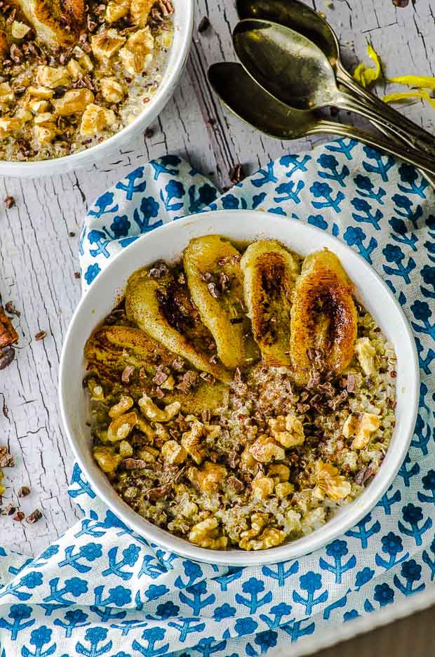 Bird's eye view of two white bowls of quinoa breakfast topped with caramelized bananas, walnuts, cocoa nibs and a sprinkle of cinnamon. There is only a partial view of the top bowl. The bottom bowl is placed on a white and blue napkin with a flower print. On the right there are 3 spoons.