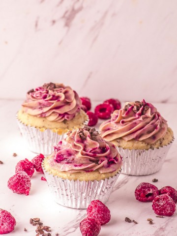 Picture of three raspberry cupcakes, with some raspberries and cacao nibs scattered around
