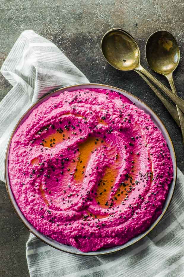 Bird's eye view of the roasted beet hummus on a plate over a white and gray tripped cloth napkin, with three spoons on the side.