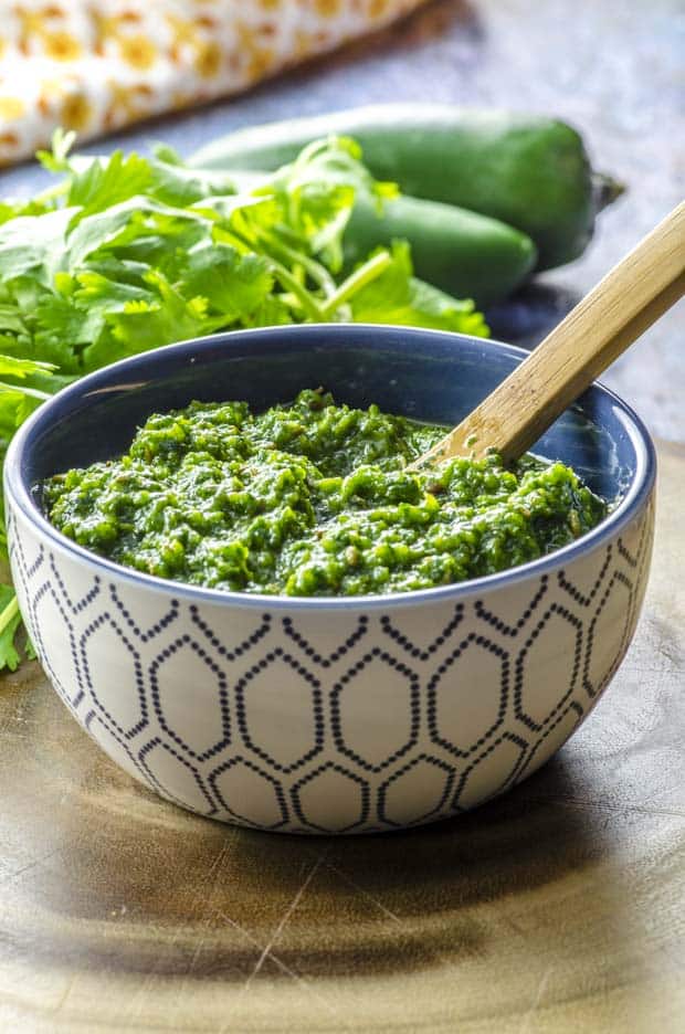 Close up of schug in a patterned white and blue bowl with a wooden spoon and some jalapenos and cilantro in the background