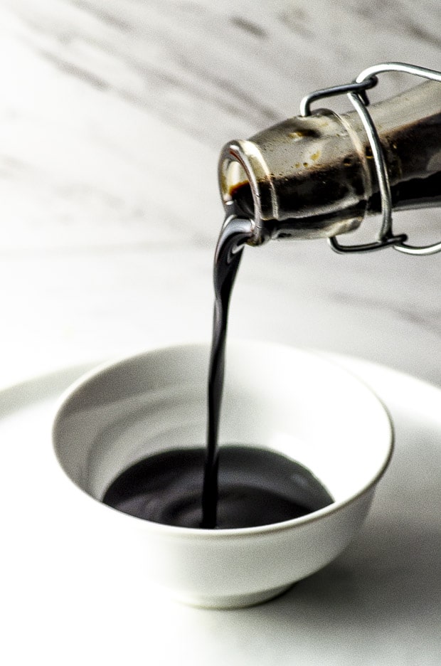 A glass bottle filled with homemade maple balsamic glaze pouring on a small white bowl
