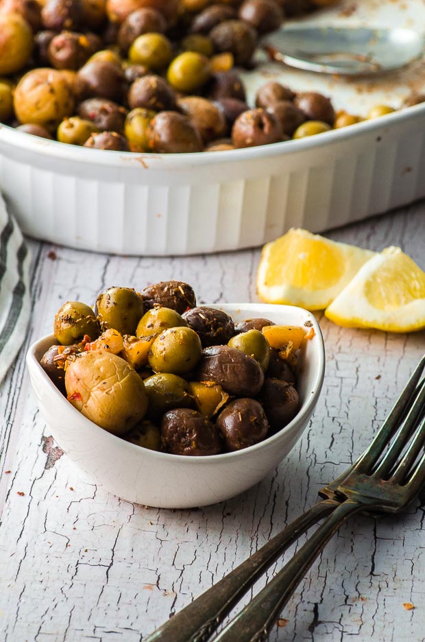 Side View of a small bowl with roasted olives and a large baking dish on the background with more roasted olives. One of our vegetarian Passover recipes.
