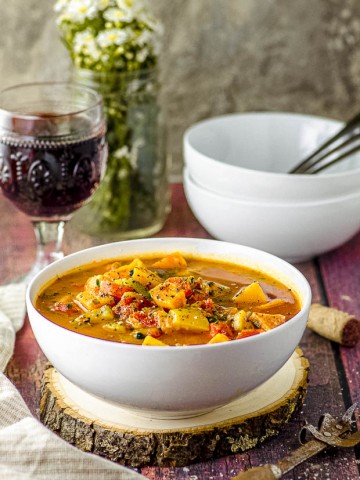side view of a white bowl filled with vegetable soup with a glass of red wine in the background