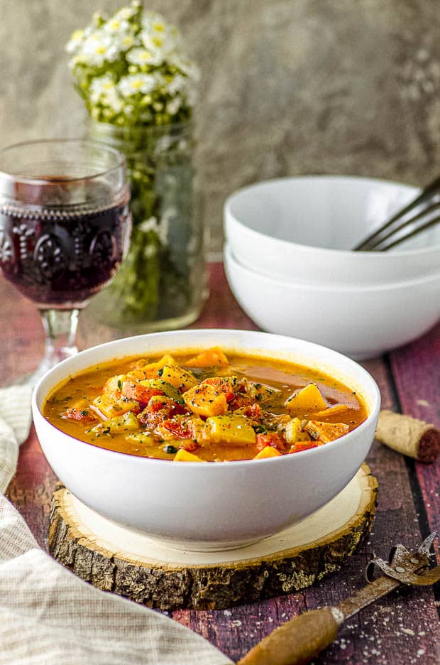 side view of a white bowl filled with vegetable soup with a glass of red wine in the background