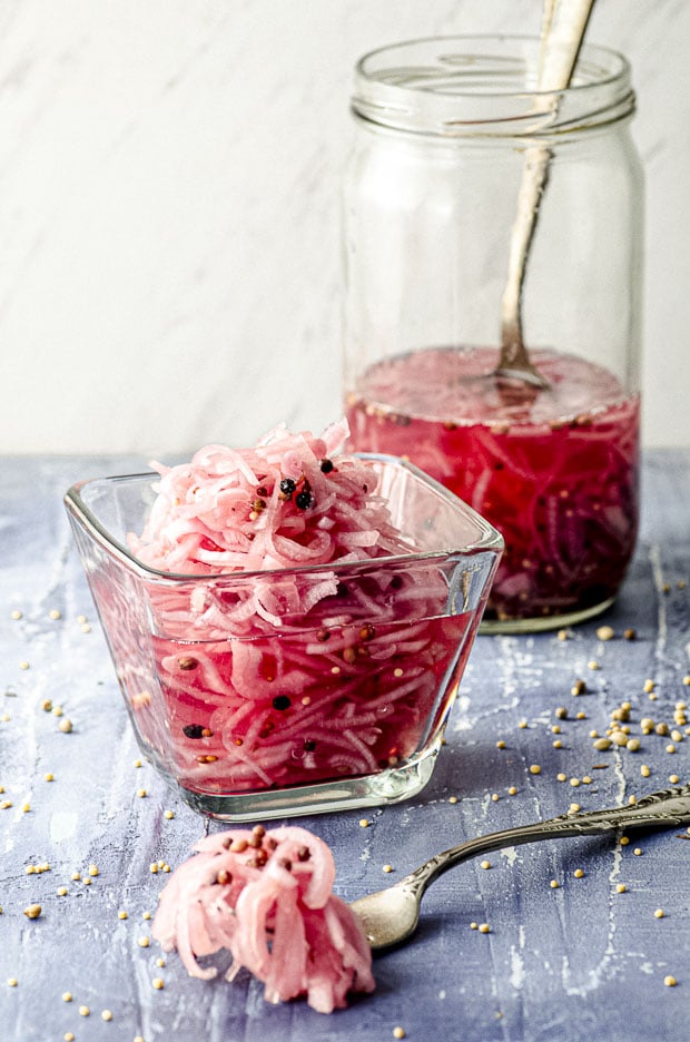 A Clear bowl filled with pickled onions and a jar of pickled onions in the background. A fork with some pickled onions on a blue surface.