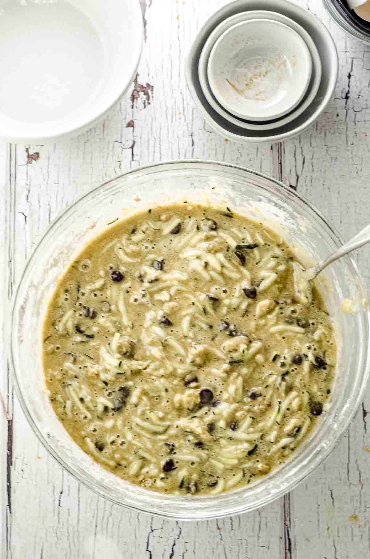A bowl with shredded Zucchini, eggs, oil , sugar and spices and a bowl with flour, chocolate chips and walnuts