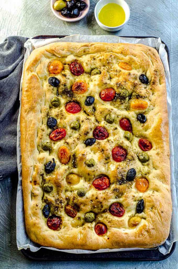 An overhead view of a whole focaccia bread with two little bowls of olive oil and olives