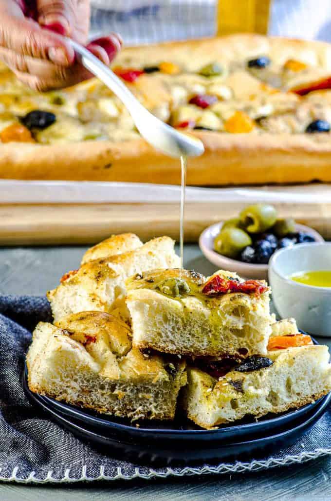 An angled view of a plate of focaccia bread cut into square pieces being drizzled with olive oil