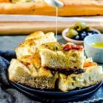 An angled view of a plate of focaccia bread cut into square pieces being drizzled with olive oil