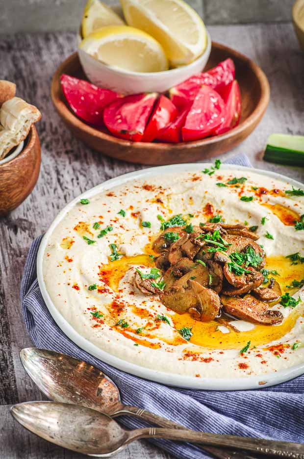 Side view of hummus with a plate of sliced tomatoes and lemon wedges on the background