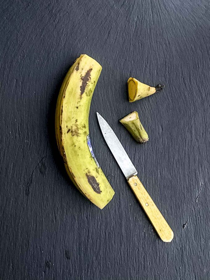 A green plantain with the ends cut off on a black surface next to a knife with a yellow handle