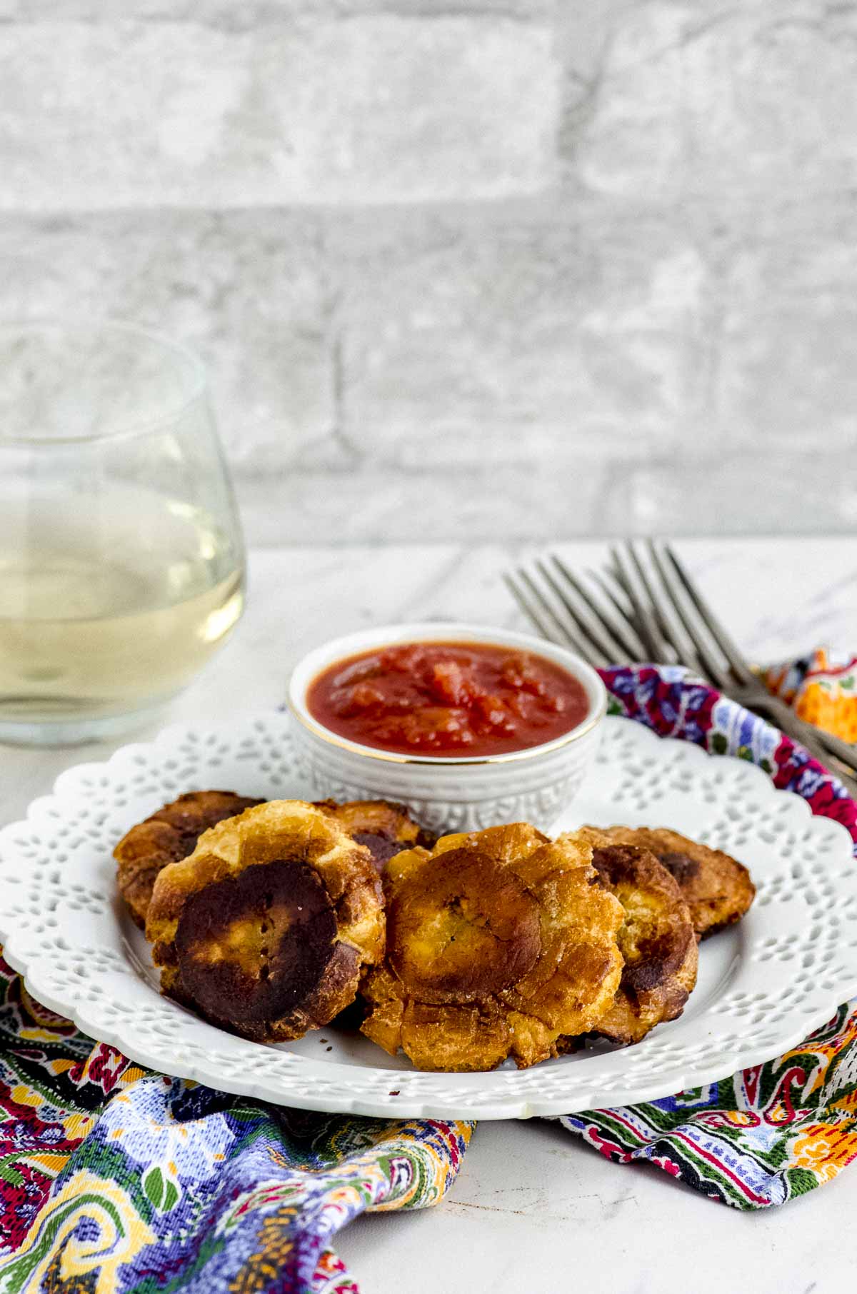 Side view of a white plate with tostones and a small bowl with red salsa on top of a colorful napkin