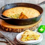 A slice of cornbread on a plate with a skillet with cornbread in the background
