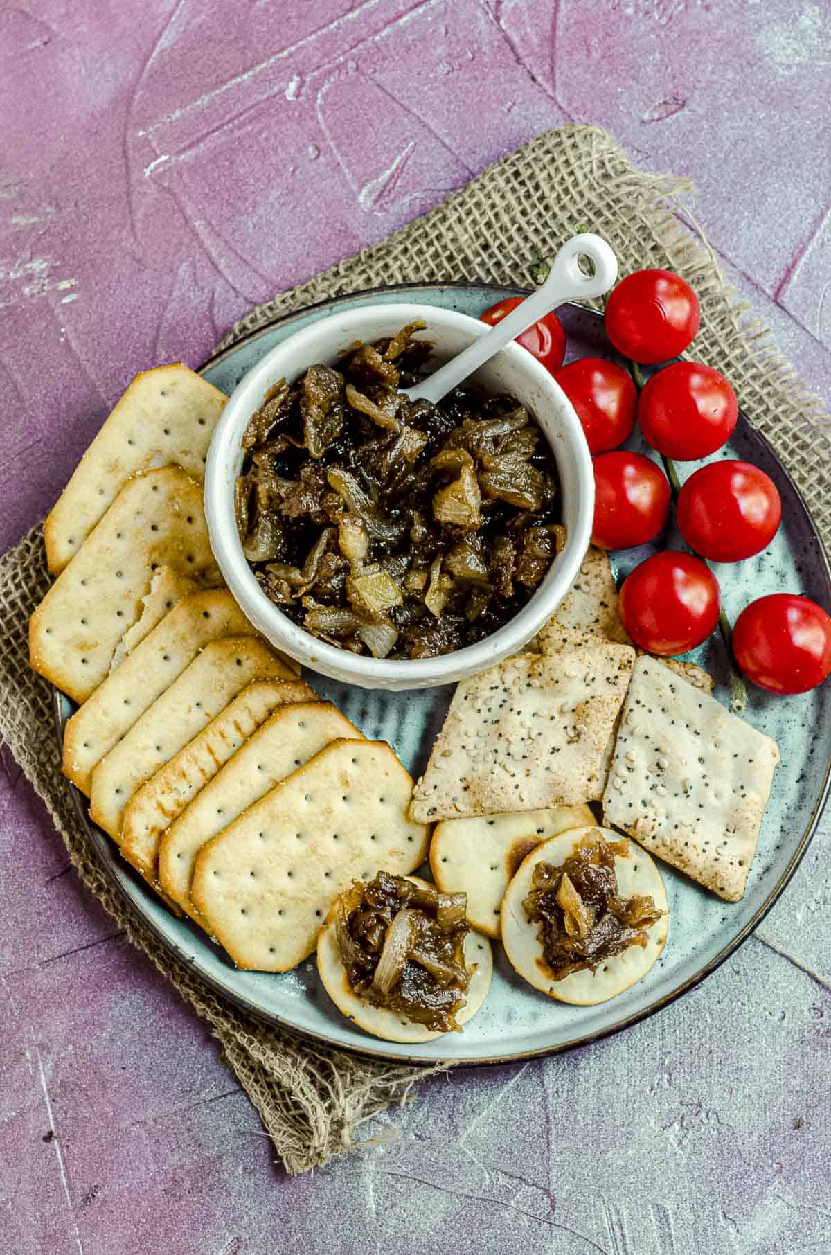 Overhead view of a bowl with apple onion chutney on a plate with some crackers and some cherry tomatoes