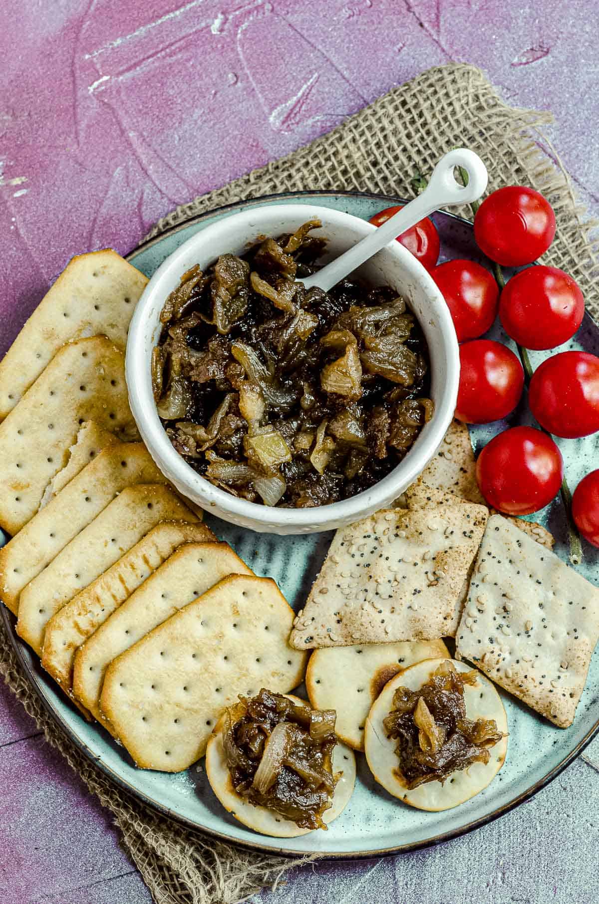 Close up view of a bowl with apple onion chutney on a plate with some crackers and some cherry tomatoes