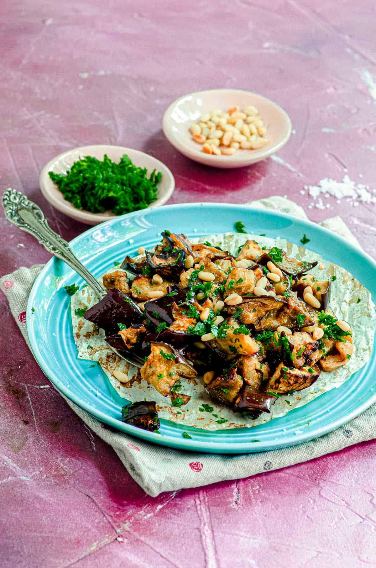 A light blue plate with roasted eggplant and a fork with two small white bowls of pine nuts and cilantro