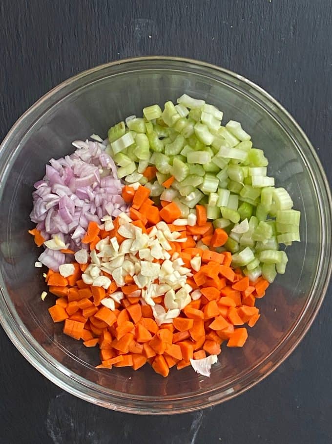 An overhead view of a glass mixing bowl with chopped onion, celery, carrots and minced garlic