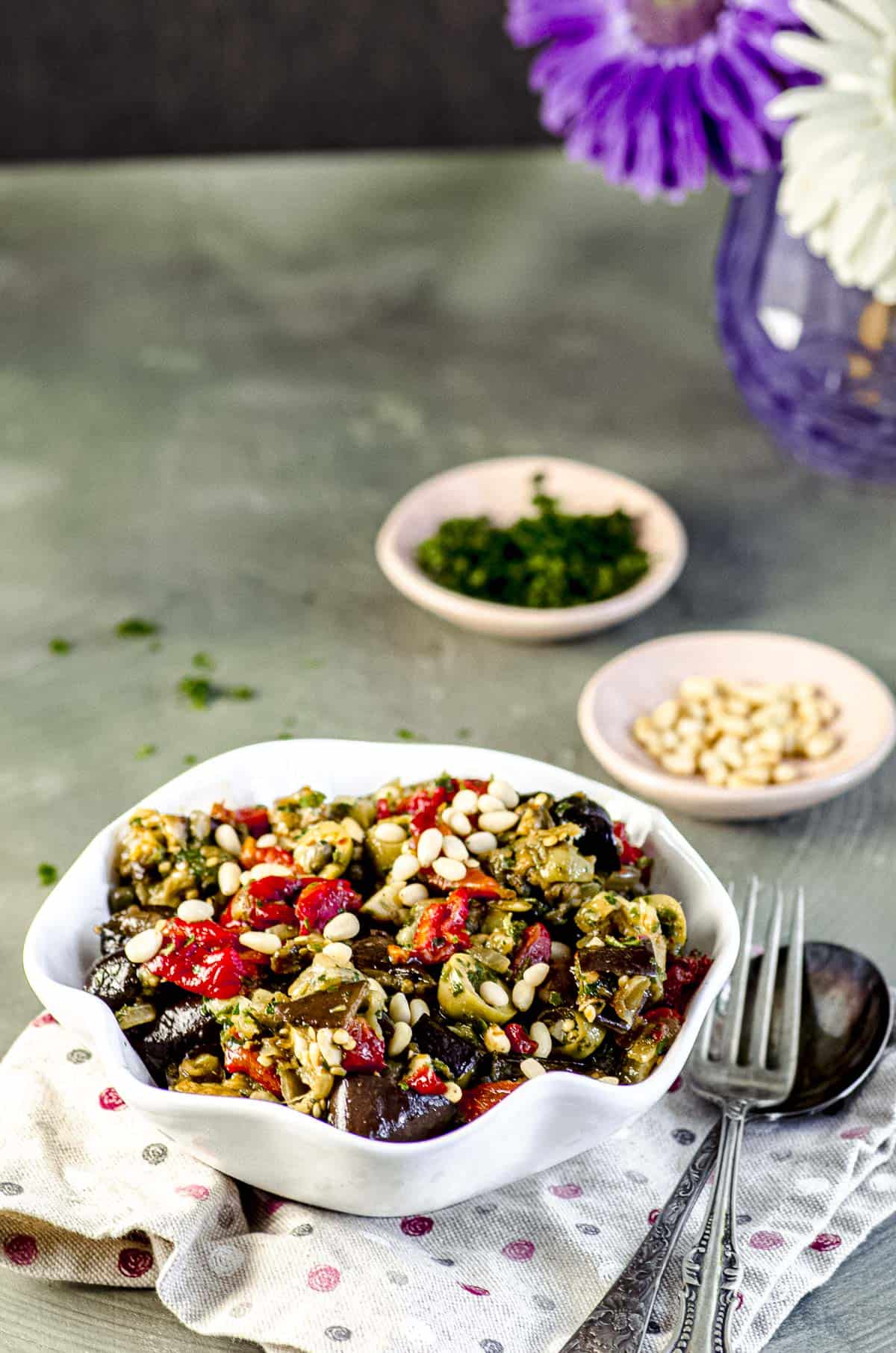 A side view of a white bowl with eggplant caponata with chopped parsley and pine nuts in separate bowls