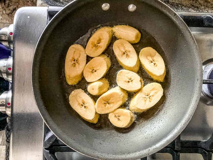An overhead view of sliced plantains in coconut oil on a skillet