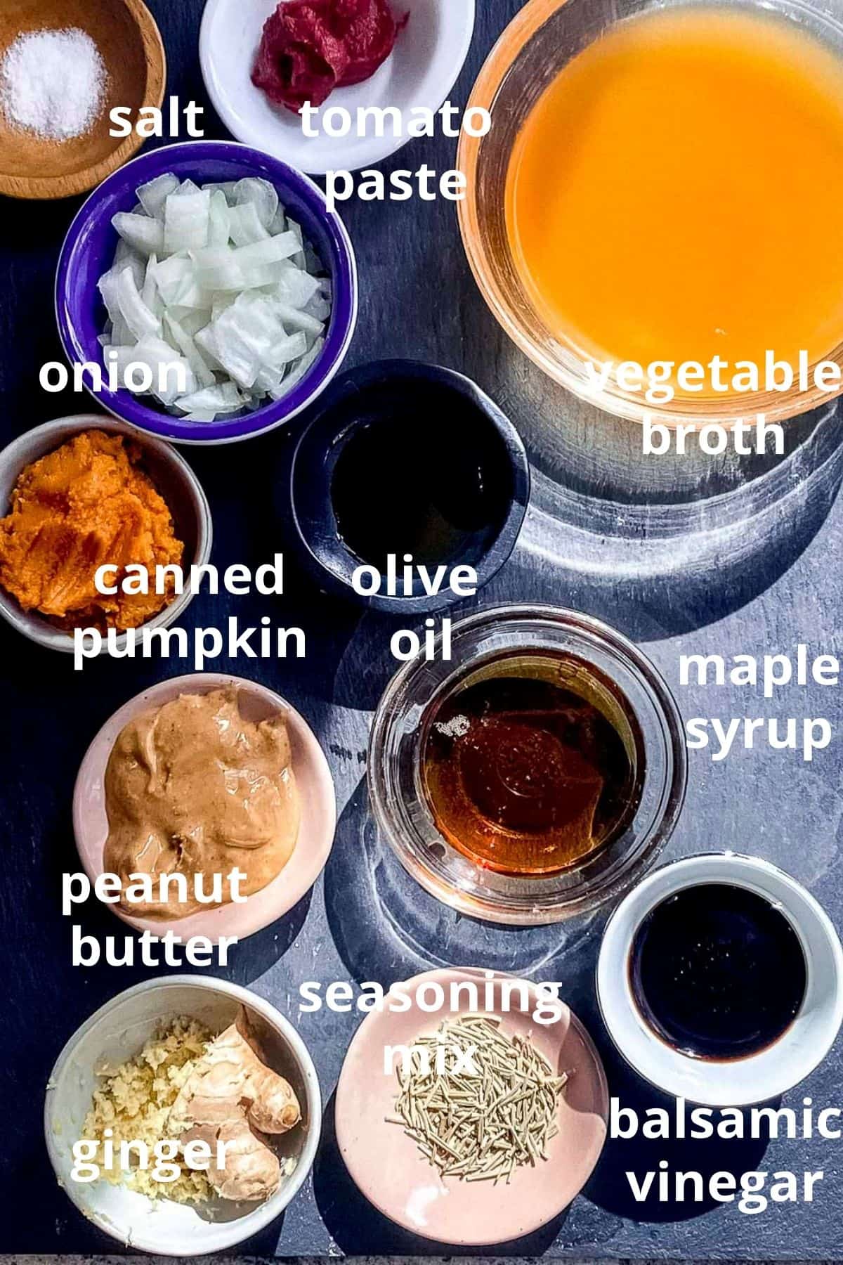 An overhead view of ingredients to make pumpkin sauce displayed in bowls; salt, tomato paste, vegetable broth, diced onion, canned pumpkin, olive oil, peanut butter, maple syrup, balsamic vinegar, seasoning mix, ginger