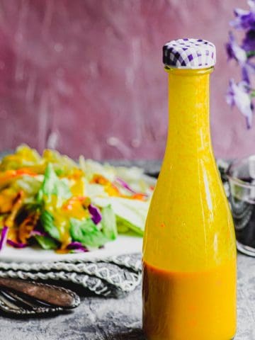A glass bottle with carrot ginger dressing in it with a salad in the background