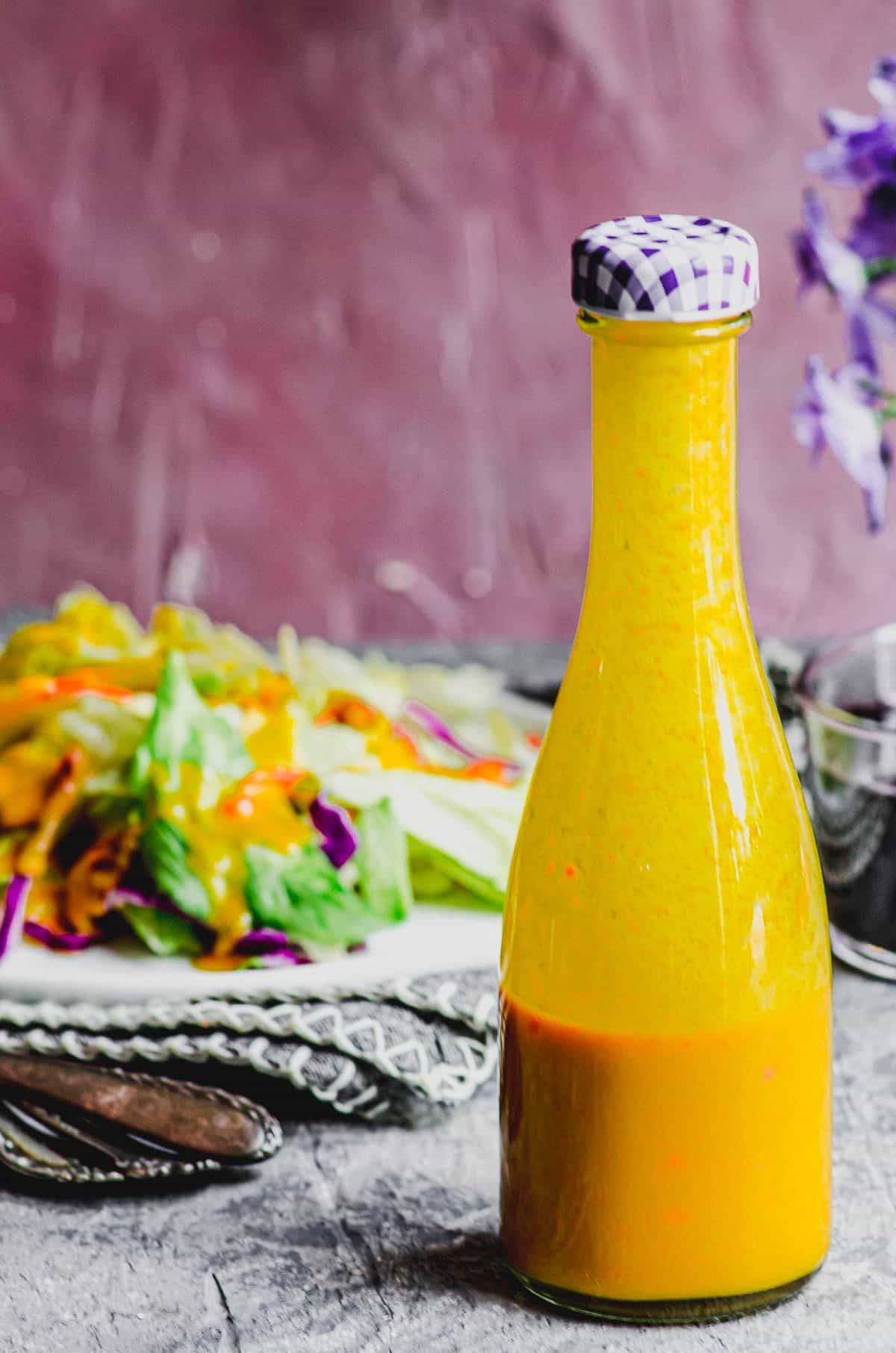 A glass bottle with carrot ginger dressing in it with a salad in the background