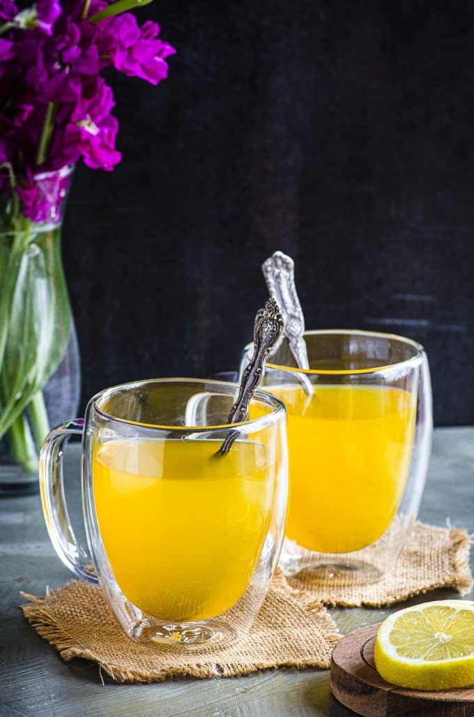 Two clear glass mugs filled with ginger turmeric tea with a spoon in each one
