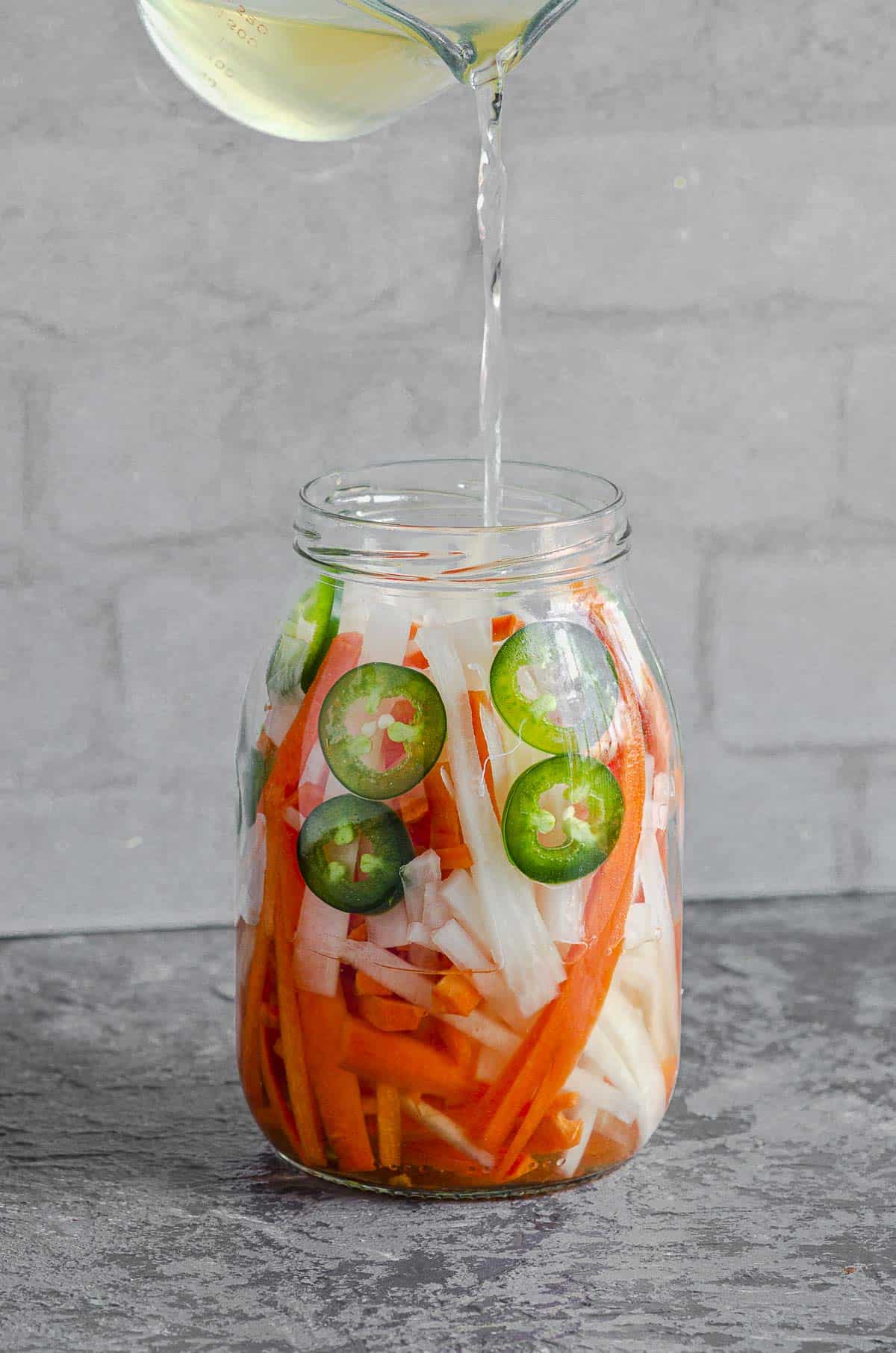 A jar of jalapeno, daikon and carrots with vinegar being poured on top