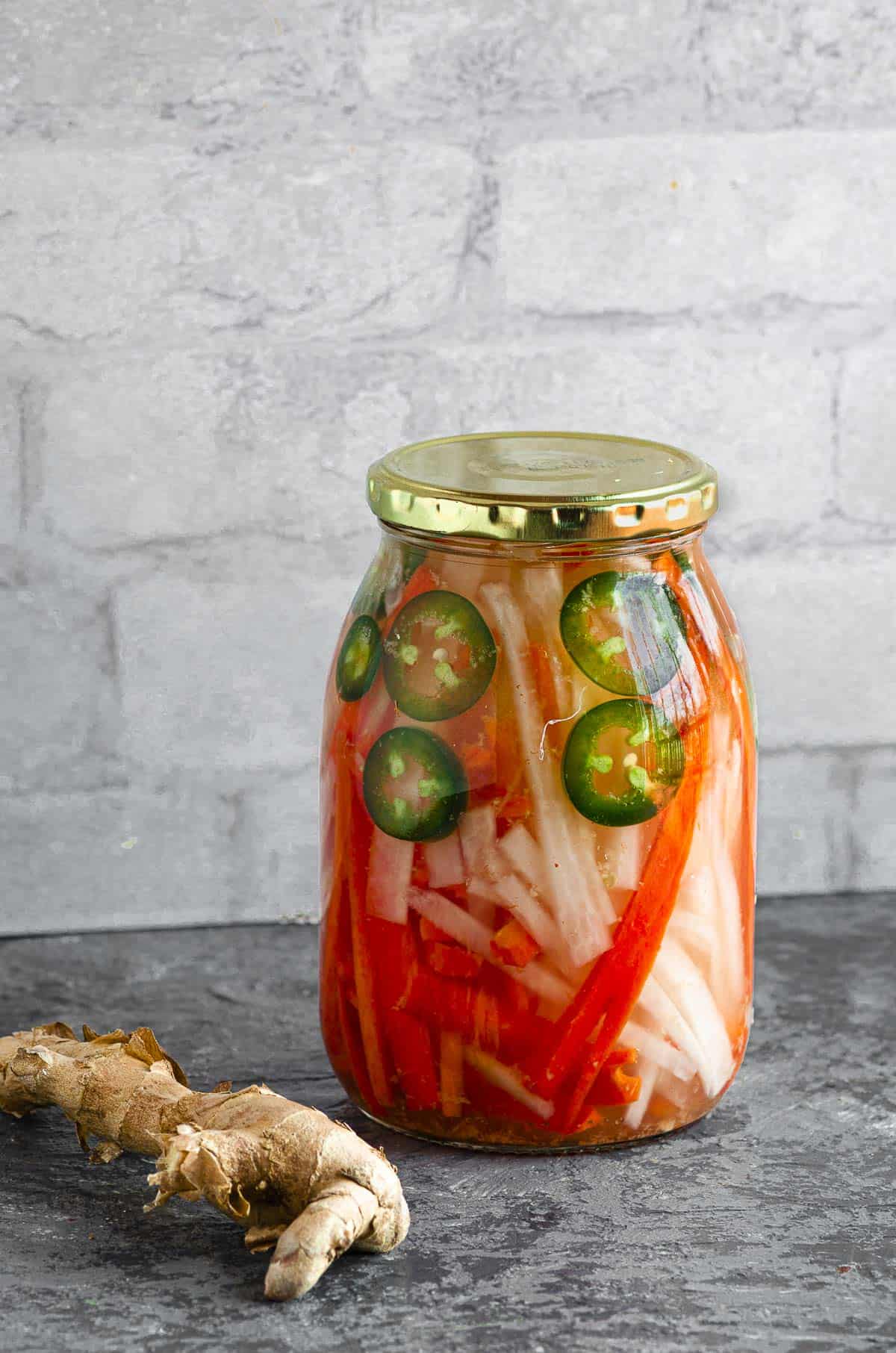 A jar of pickled daikon, carrots and jalapeno with ginger sitting next to it